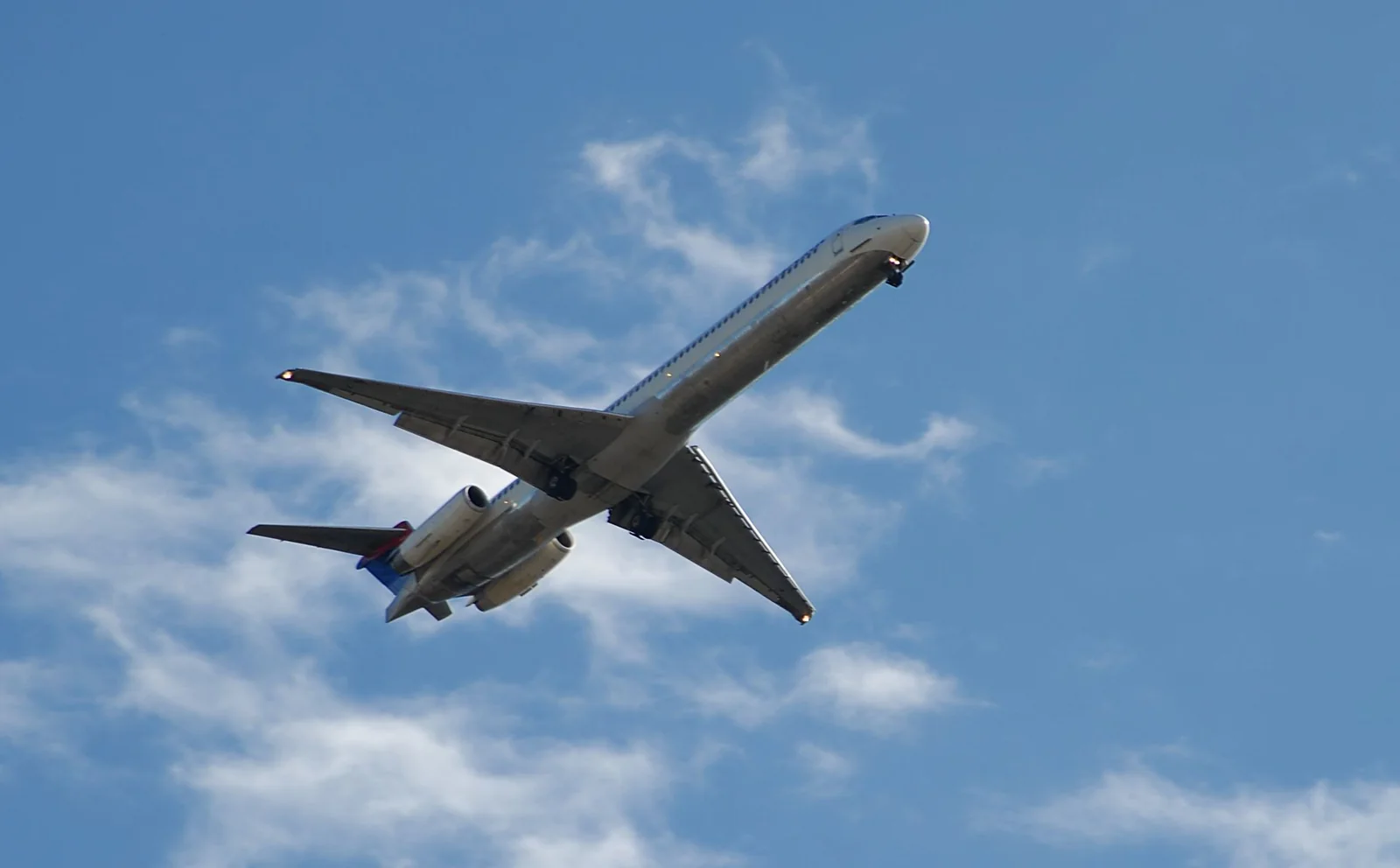 A twin-engined airliner seen from below, climbing through a pale blue sky with light cloud